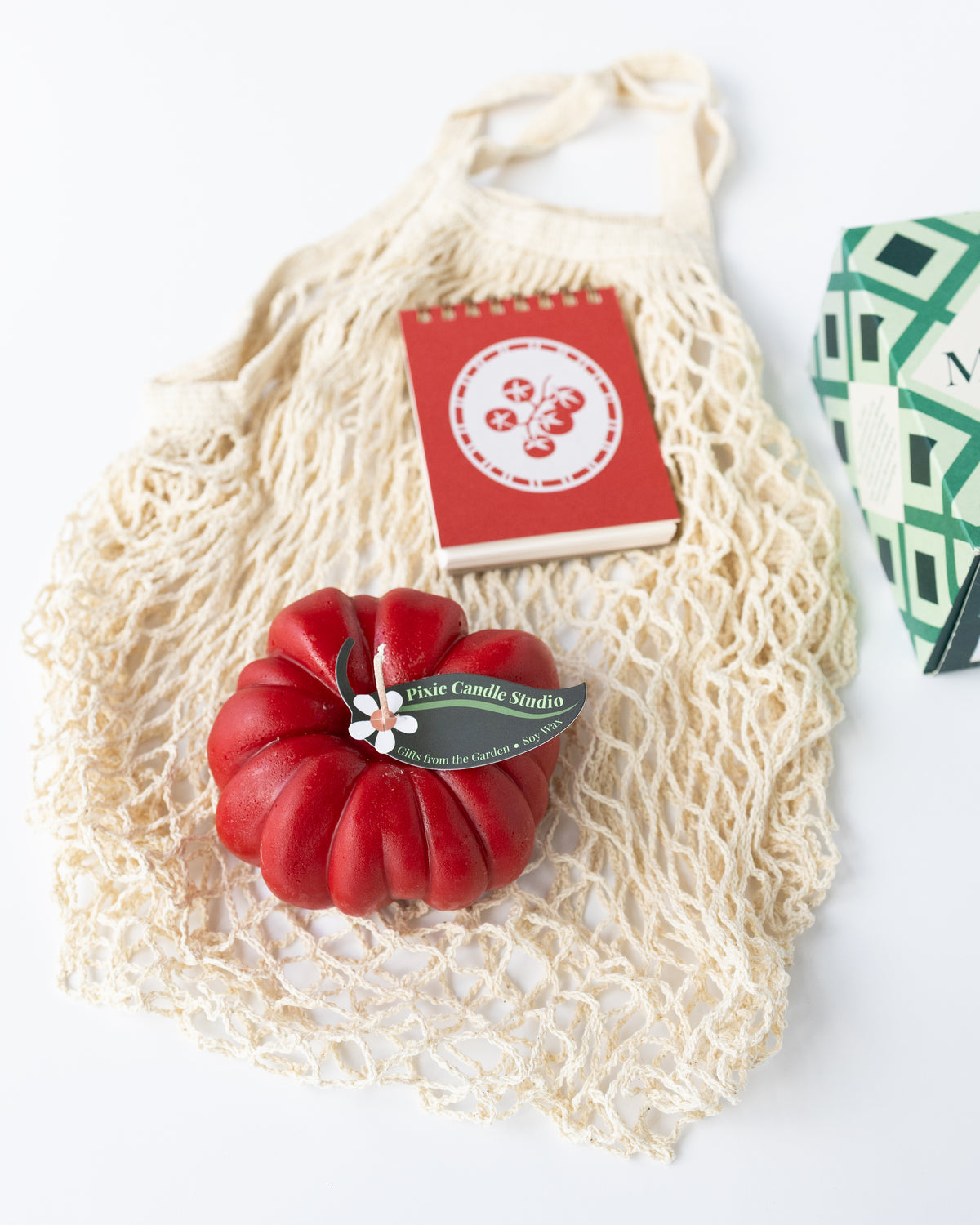 An angled view of a Farmer's Market-themed gift with a mesh net bag, a tomato-shaped red candle, a red mini notebook with a tomato graphic on top, and a box of Maldon salt just out of frame. 