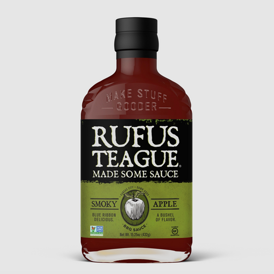 A bottle of Rufus Teague Smokey Apple BBQ Sauce on a light gray background. The bottle is made of dark amber glass with a black cap and a black and olive green label featuring a white apple illustration.