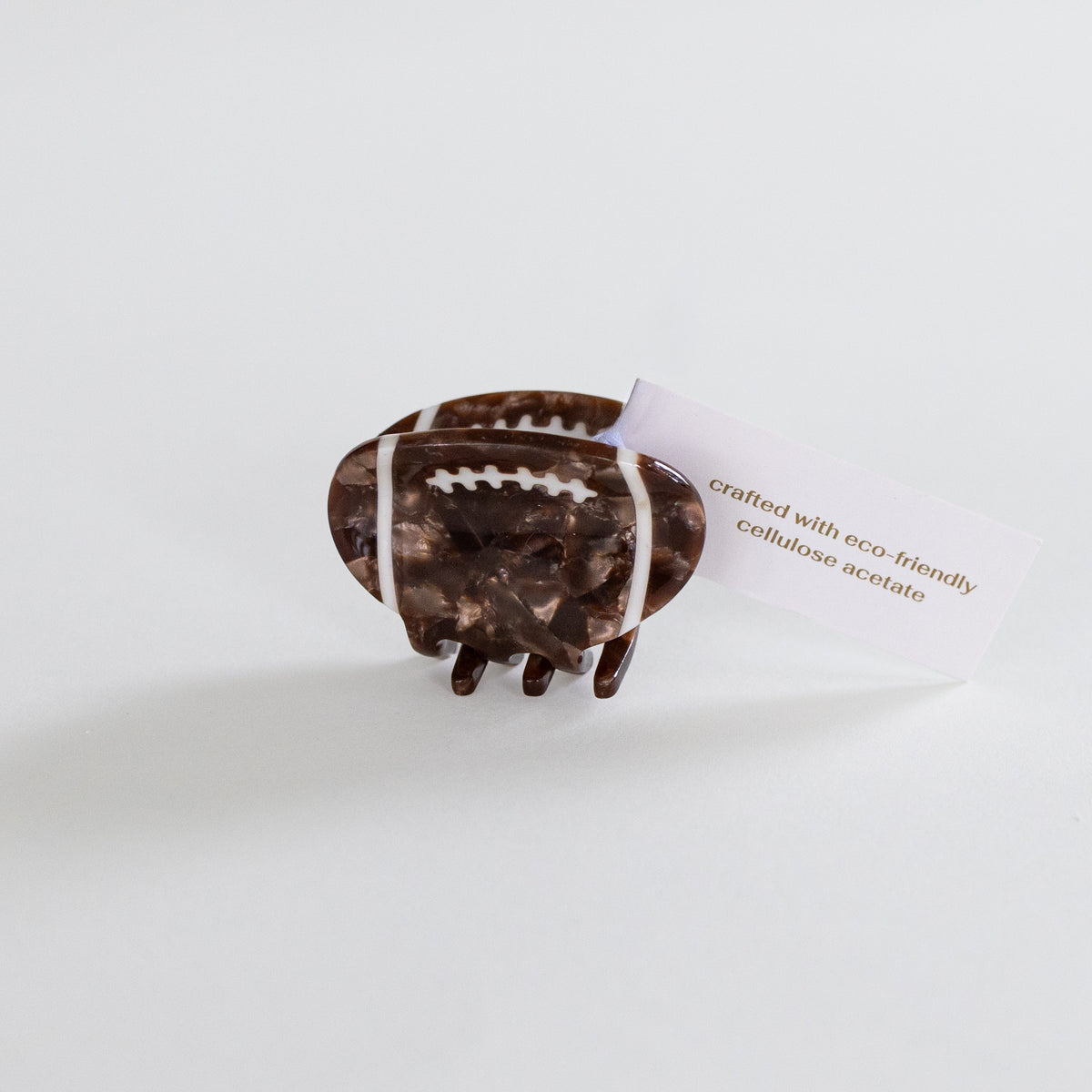 A small hair claw clip shaped like a football, made of marbled brown and clear acetate with white laces, shown on a light gray background with a small product tag attached.