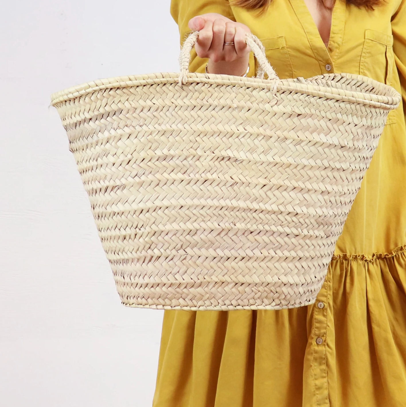 A woman in a long yellow dress holds a woven market basket tote bag with woven handles.