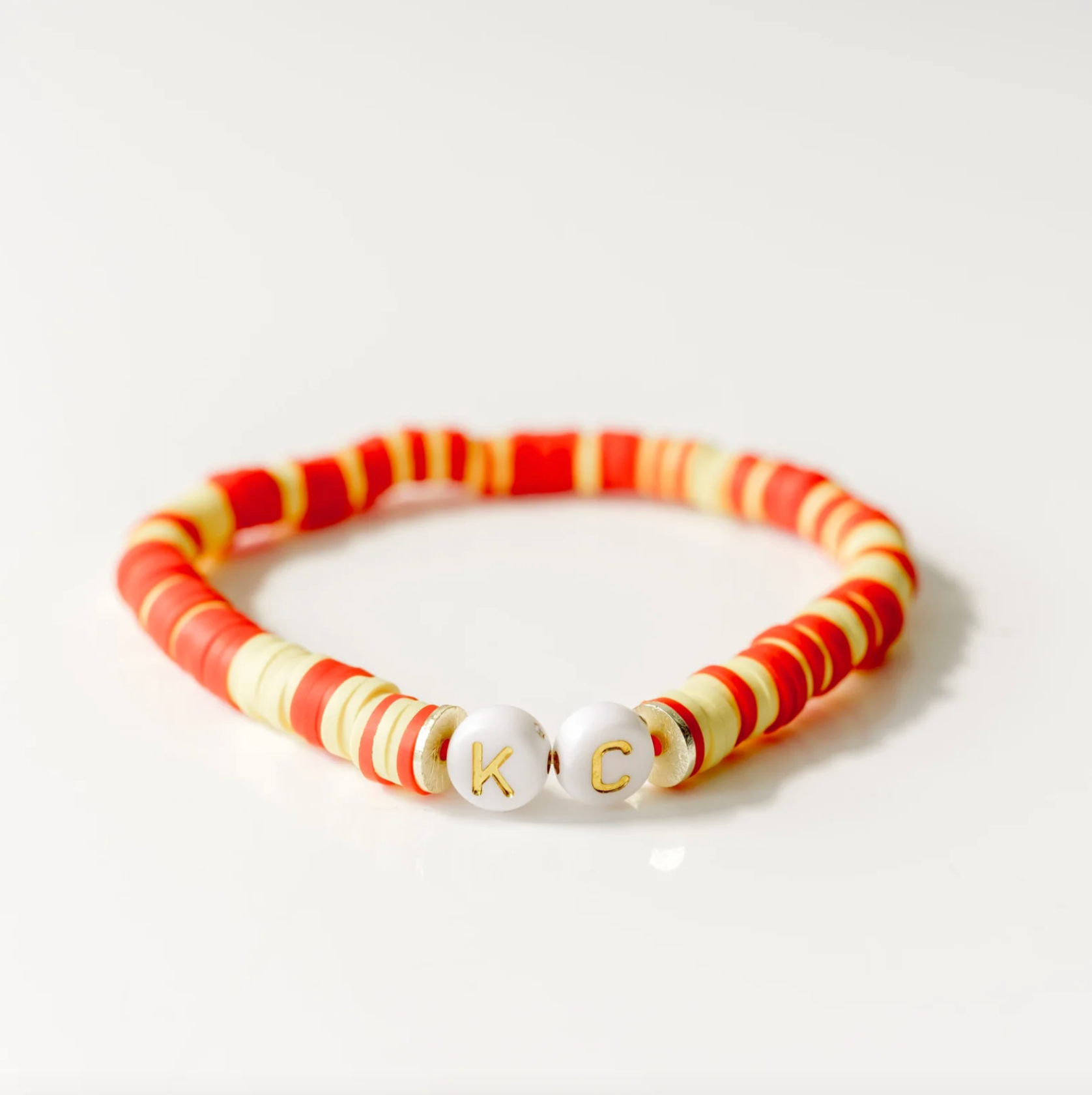 A beaded stretch bracelet with alternating red and cream cylindrical beads, featuring two round white beads at the center with the gold-embossed letters 'K' and 'C'.