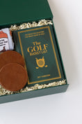 Load image into Gallery viewer, A curated golf gift set in a dark green box filled with shredded paper, featuring a rolled white kitchen towel with whiskey bottle graphics, a green drink sweater with 'KC' in gold, two brown leather coasters, and a green book titled 'The Golf Bucket List' by Jeff Thoreson.

