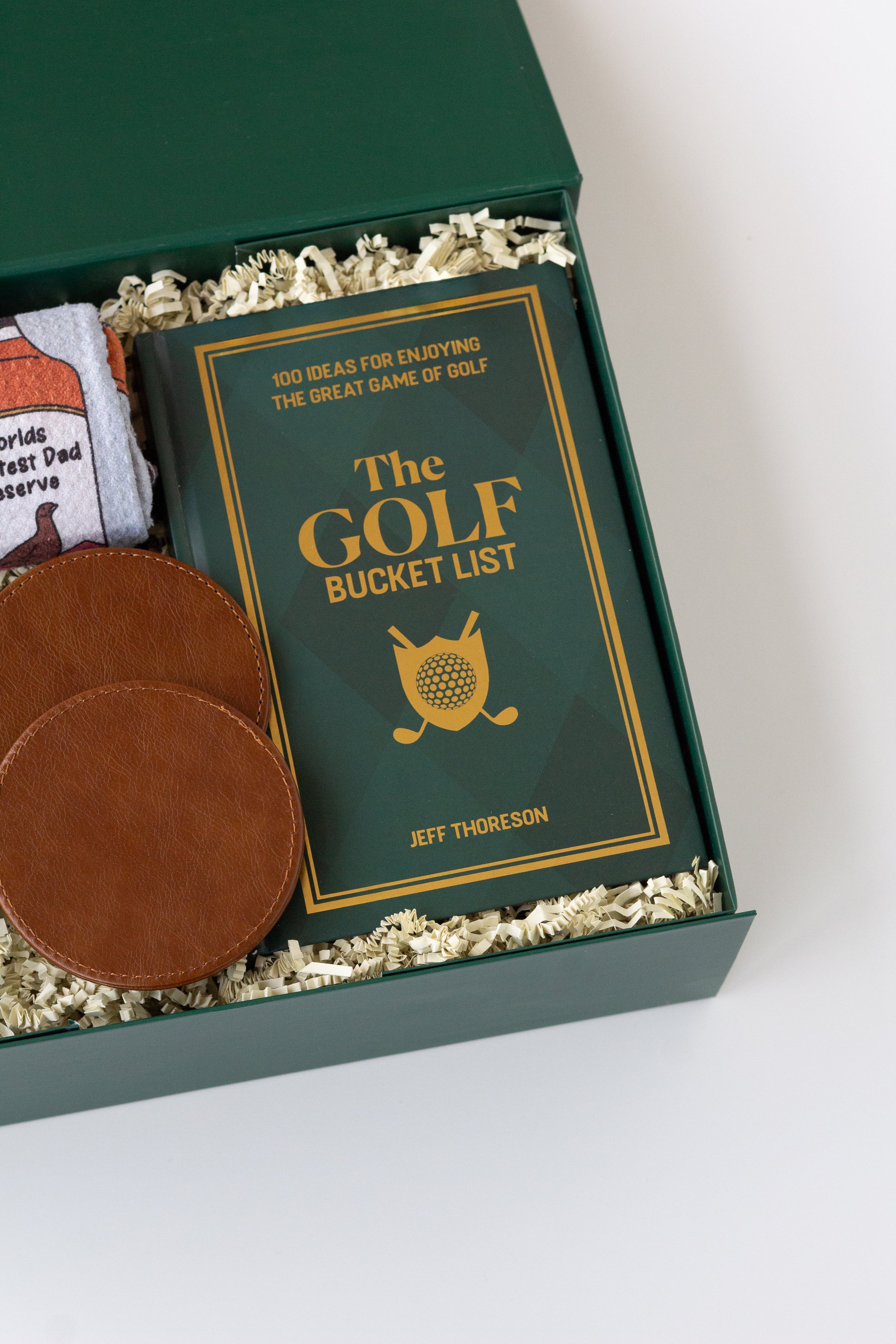 A curated golf gift set in a dark green box filled with shredded paper, featuring a rolled white kitchen towel with whiskey bottle graphics, a green drink sweater with 'KC' in gold, two brown leather coasters, and a green book titled 'The Golf Bucket List' by Jeff Thoreson.