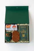 Load image into Gallery viewer, A curated golf gift set in a dark green box filled with shredded paper, featuring a rolled white kitchen towel with whiskey bottle graphics, a green drink sweater with 'KC' in gold, two brown leather coasters, and a green book titled 'The Golf Bucket List' by Jeff Thoreson.
