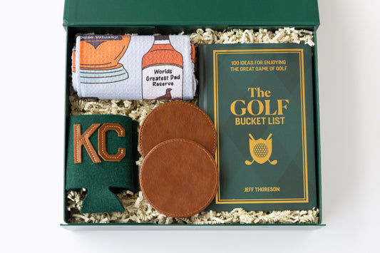 A curated golf gift set in a dark green box filled with shredded paper, featuring a rolled white kitchen towel with whiskey bottle graphics, a green drink sweater with 'KC' in gold, two brown leather coasters, and a green book titled 'The Golf Bucket List' by Jeff Thoreson.