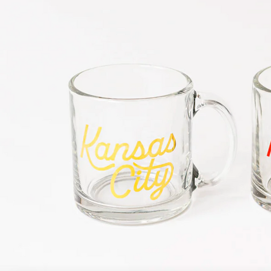 A clear glass mug with 'Kansas City' written in gold script on the front, shown on a white background with part of another mug visible on the right edge.