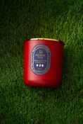 Load image into Gallery viewer, A red wax candle in a matte red glass jar with a gold metal lid, placed on a green grass background. The front label reads 'GLOBAL CUP KANSAS CITY 2026' 
