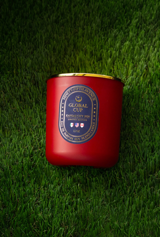 A red wax candle in a matte red glass jar with a gold metal lid, placed on a green grass background. The front label reads 'GLOBAL CUP KANSAS CITY 2026' 