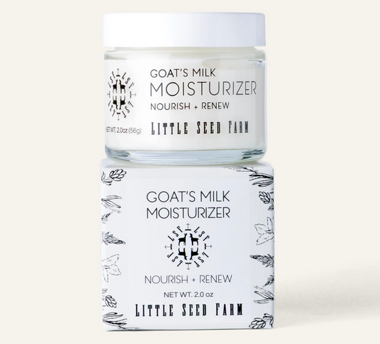 A jar of Goat's Milk Moisturizer by Little Seed Farm sits on top of its matching white box. The packaging features black text and delicate botanical line drawings on a white background, set against a solid light beige backdrop.