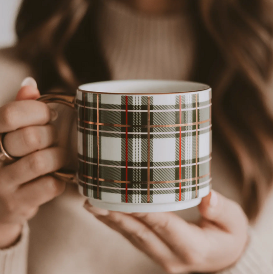 A person holding a white ceramic mug with a green, gold, and red plaid pattern, wearing a beige knit sweater