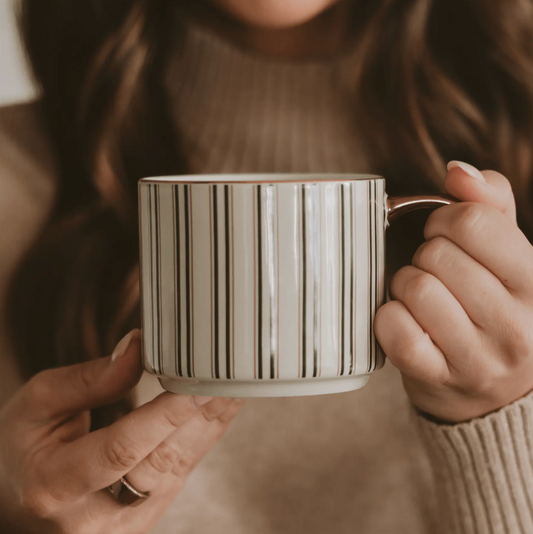A close-up of a person holding a ceramic mug with vertical green stripes on a white background. The person is wearing a ribbed beige sweater, and the mug has a metallic rose gold handle.