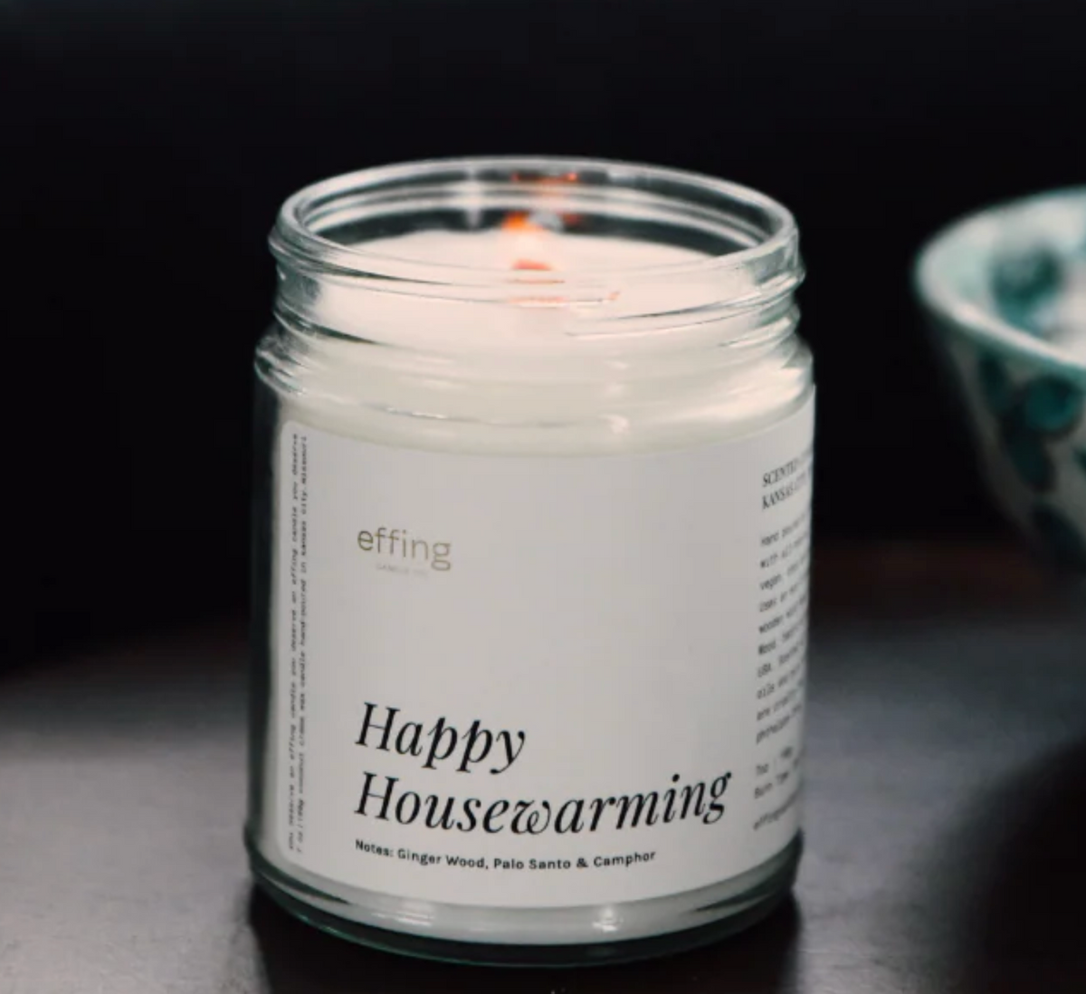A white jar candle titled "Happy Housewarming" sitting on a dark tabletop with a bowl behind it. 