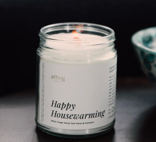 A white jar candle titled "Happy Housewarming" sitting on a dark tabletop with a bowl behind it. 