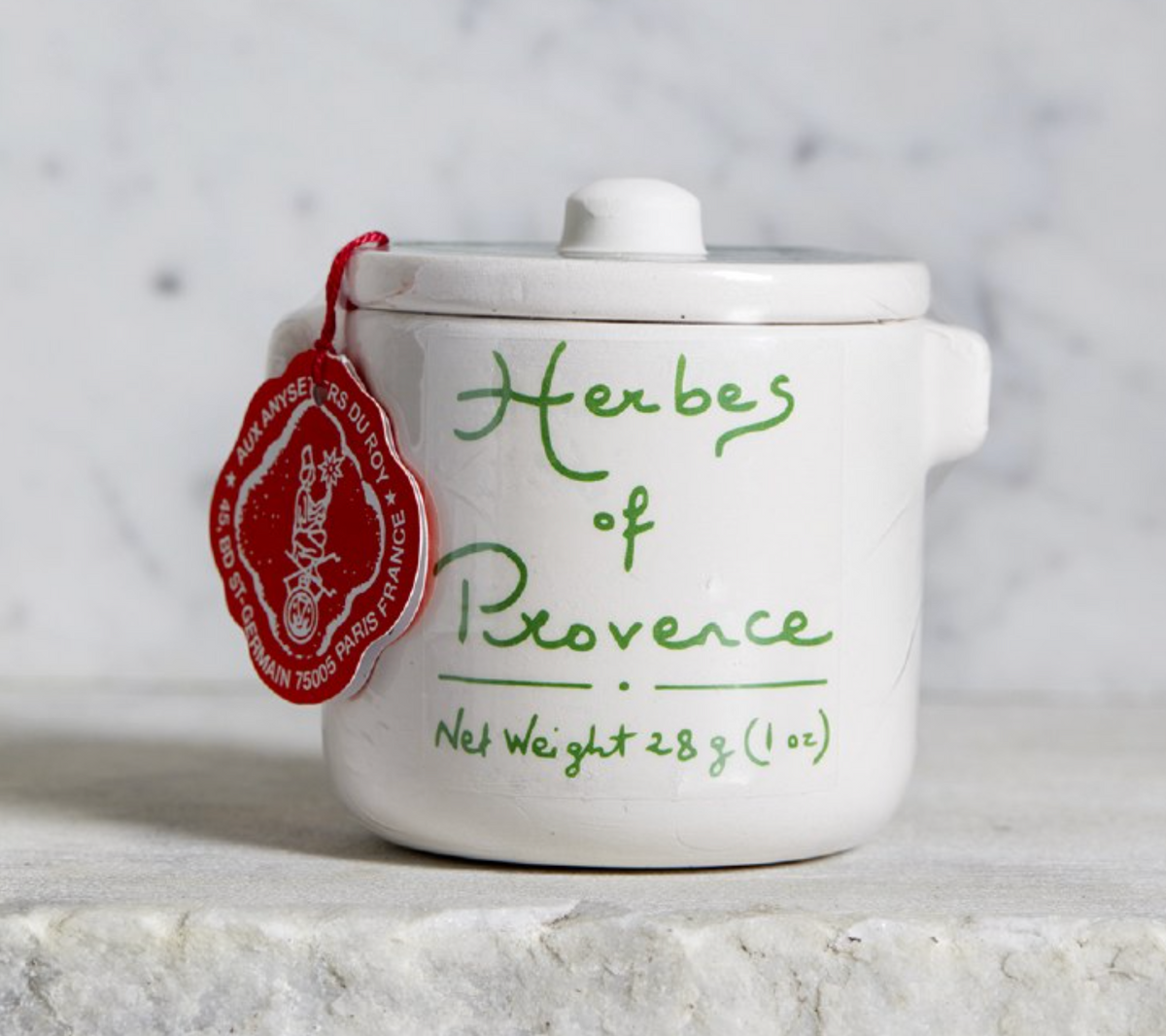 A small white ceramic jar with a lid, decorated with green cursive text reading 'Herbs of Provence' and a red hanging tag, set on a light stone surface against a soft marble-like background.