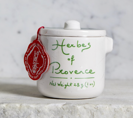 A small white ceramic jar with a lid, decorated with green cursive text reading 'Herbs of Provence' and a red hanging tag, set on a light stone surface against a soft marble-like background.
