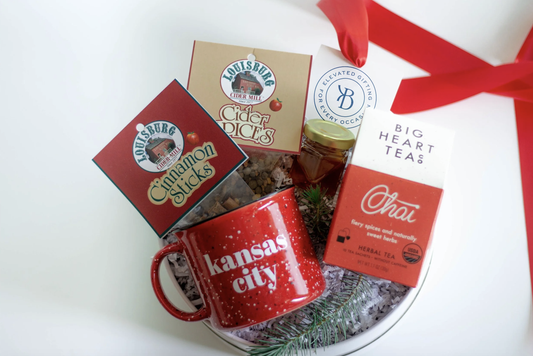 A holiday gift basket arrangement on a white surface featuring a red ceramic mug with 'Kansas City' printed on it, boxed Louisburg Cider Mill Cinnamon Sticks and Cider Spices, Big Heart Tea Chai tea, and a jar of honey, tied together with a red ribbon.