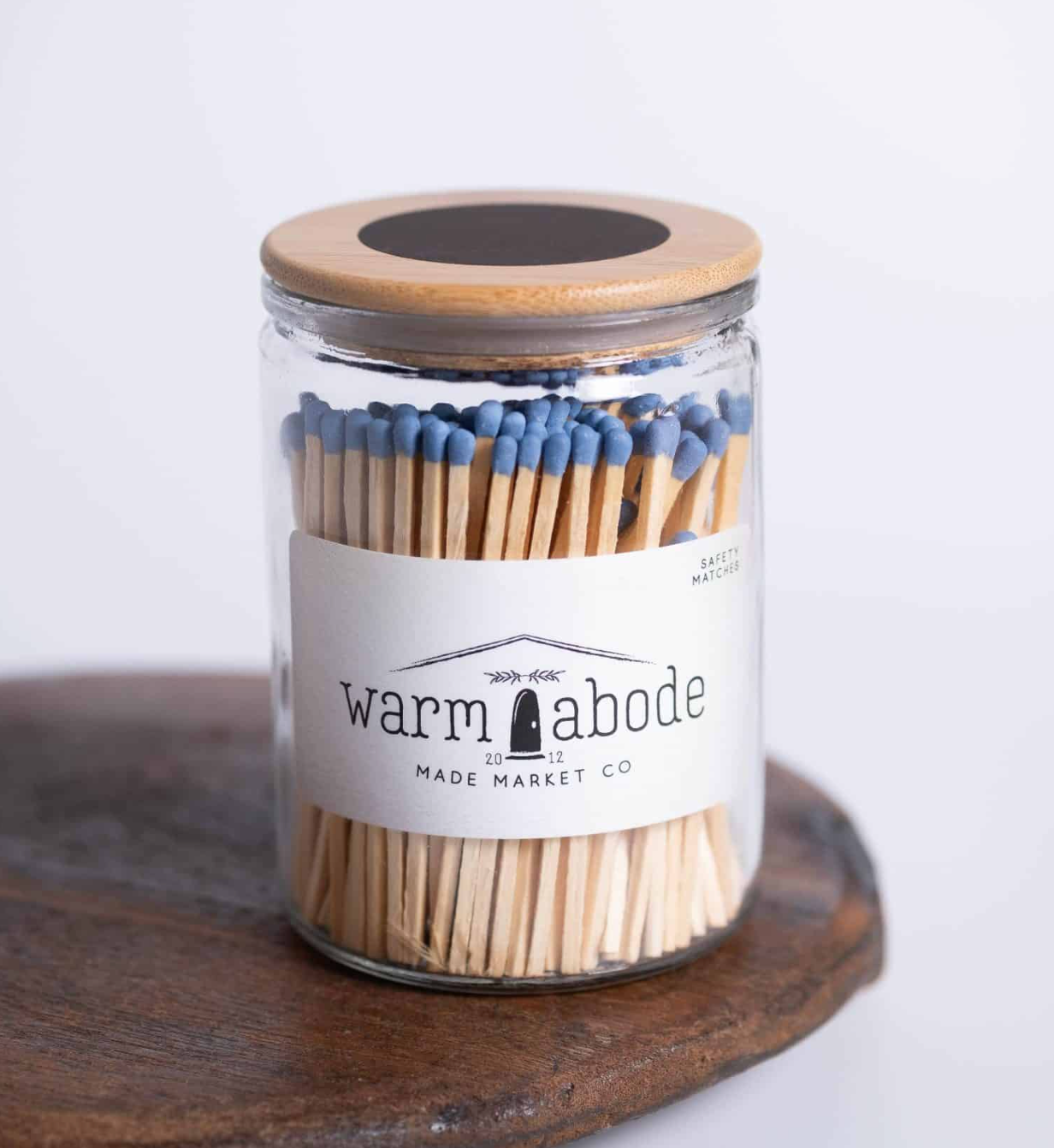 A clear glass jar filled with wooden matchsticks topped with blue tips and sealed with a light wood lid, standing on a dark wooden surface against a white background. The front label reads 'warm abode' and 'MADE MARKET CO'.
