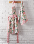 Load image into Gallery viewer, A pink floral apron and matching kitchen towel hanging from a wooden peg rail against a white painted brick wall.
