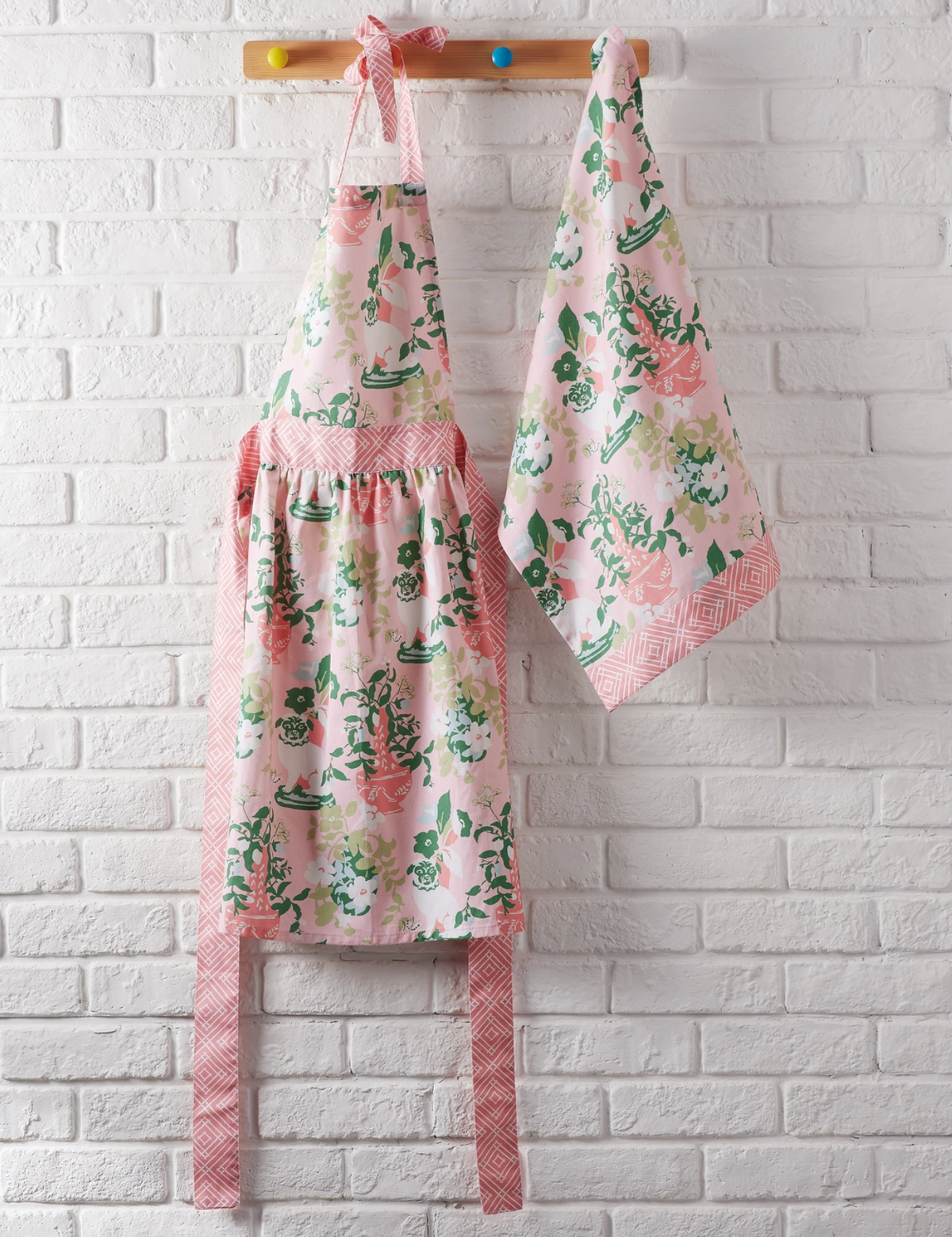 A pink floral apron and matching kitchen towel hanging from a wooden peg rail against a white painted brick wall.