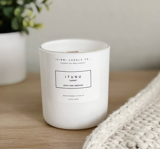 A white glass jar candle from Isimmi Candle Co. labeled 'ITUNU', placed on a light wood surface with a soft neutral background, a blurred plant in the back left, and a chunky knit blanket in the foreground right.