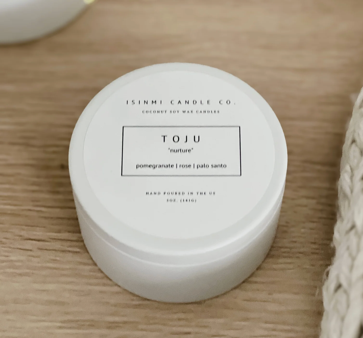 A round white jar of Isinmi Candle Co. Toju coconut soy wax candles on a light wood grain surface.