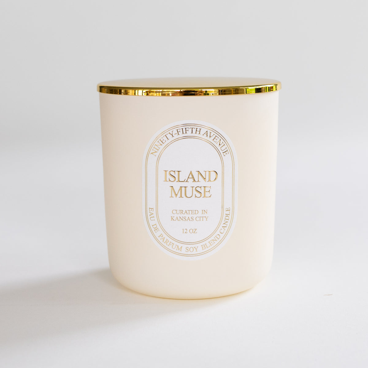 A 12 oz soy wax candle in a frosted cream-colored glass jar with a metallic gold rim and a gold foil label reading 'ISLAND MUSE' and 'Ninety-Fifth Avenue'.