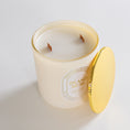 Load image into Gallery viewer, A cream-colored scented candle in a frosted glass jar with a gold rim, shown with its matching gold lid leaning against the jar on a clean white background. The label reads 'ISLAND MUSE' 
