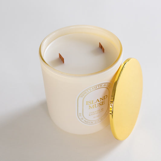 A cream-colored scented candle in a frosted glass jar with a gold rim, shown with its matching gold lid leaning against the jar on a clean white background. The label reads 'ISLAND MUSE' 