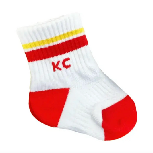 A white baby sock with red and yellow stripes near the cuff, featuring the letters 'KC' in red on the side and red sections on the toe and heel, shown against a plain white background.