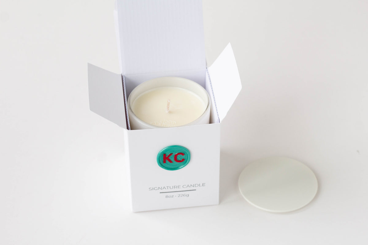A white jar candle in an open white box with the lid placed beside it on a clean white background. The box has a green circular logo with red letters and the text 'KC' and 'SIGNATURE CANDLE'.