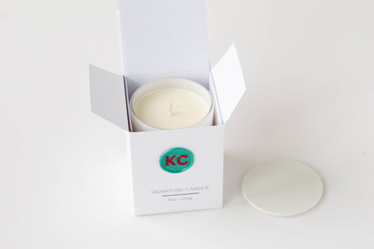 A white jar candle in an open white box with the lid placed beside it on a clean white background. The box has a green circular logo with red letters and the text 'KC' and 'SIGNATURE CANDLE'.