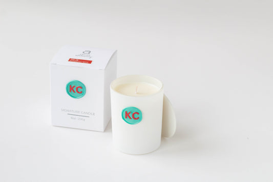 A white ceramic candle jar with a teal circular KC logo wax seal, shown next to its white packaging box on a clean white background.