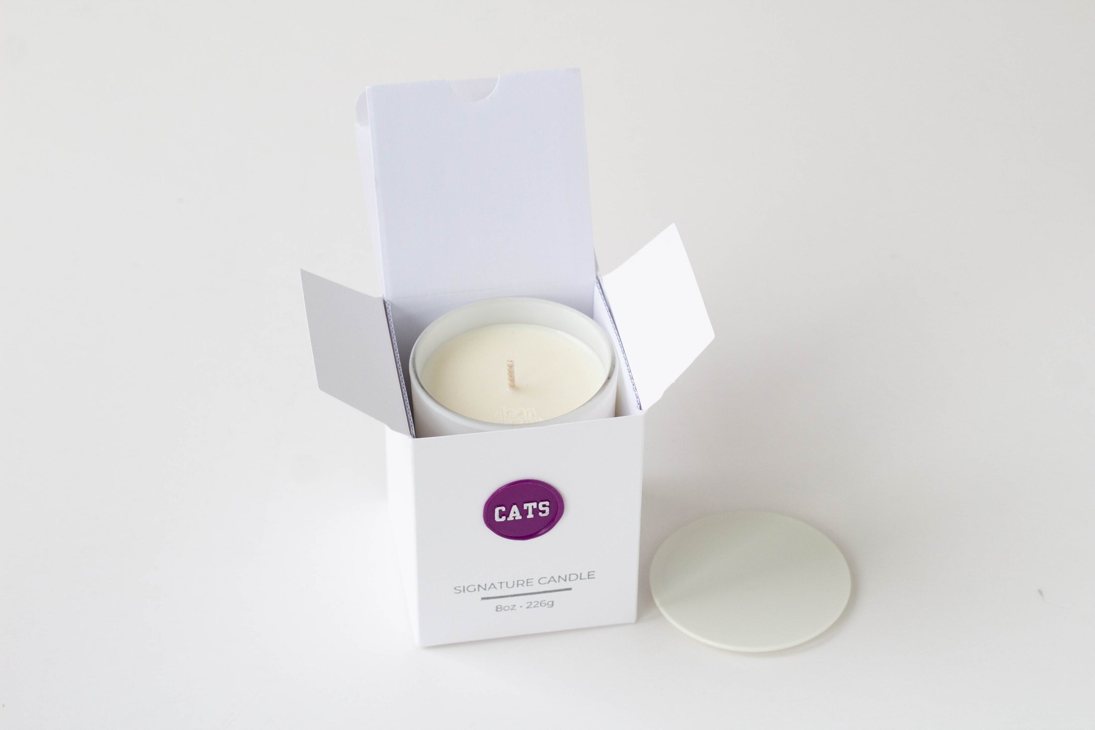 A white wax candle in a white open box with a purple circular logo reading CATS, shown on a clean white background with the lid placed beside the box.