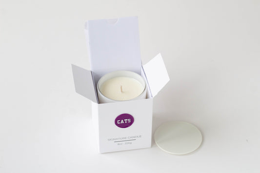 A white wax candle in a white open box with a purple circular logo reading CATS, shown on a clean white background with the lid placed beside the box.