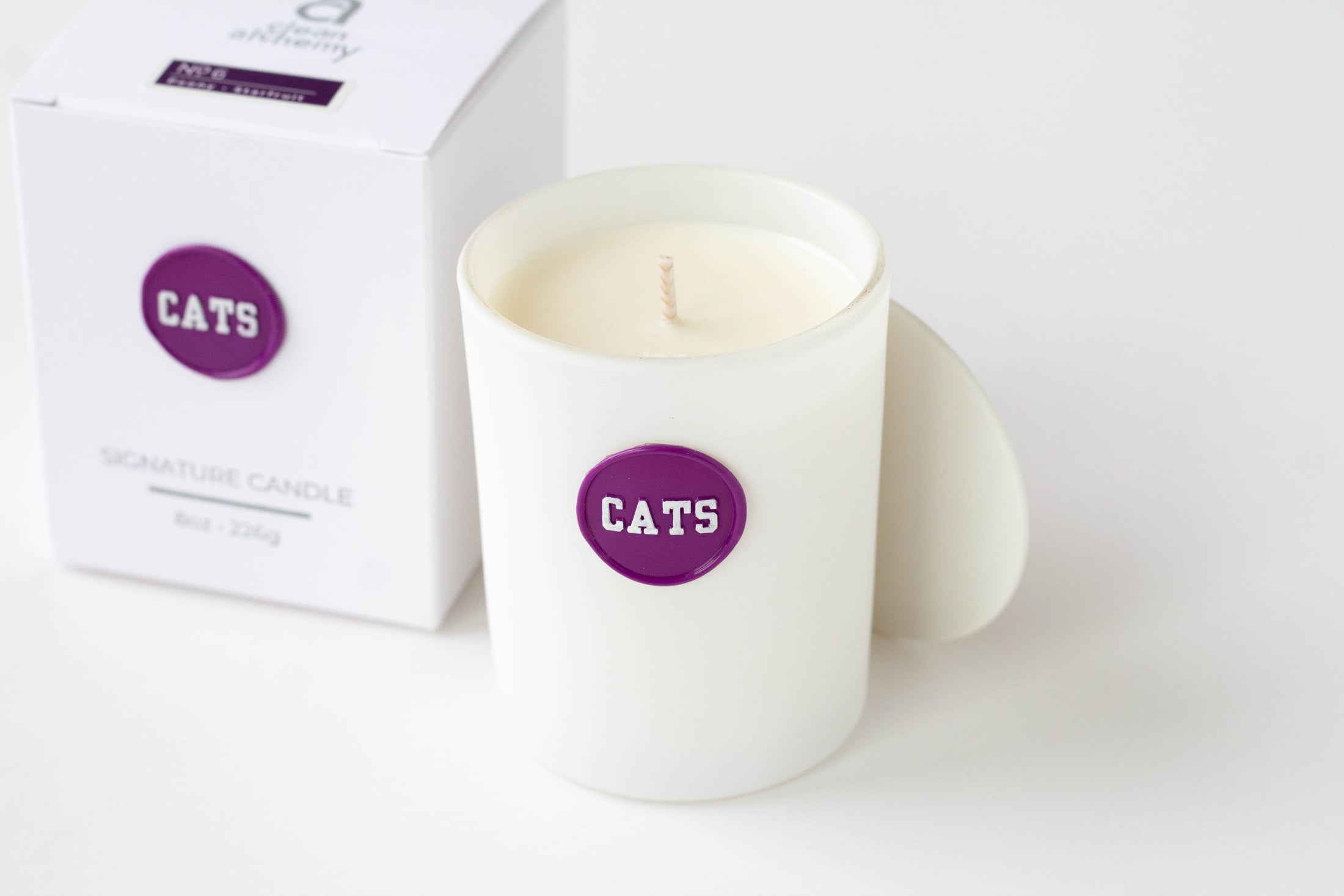 A white wax candle in a white glass jar with a purple circular label reading 'CATS', shown next to its white box packaging on a clean white background.