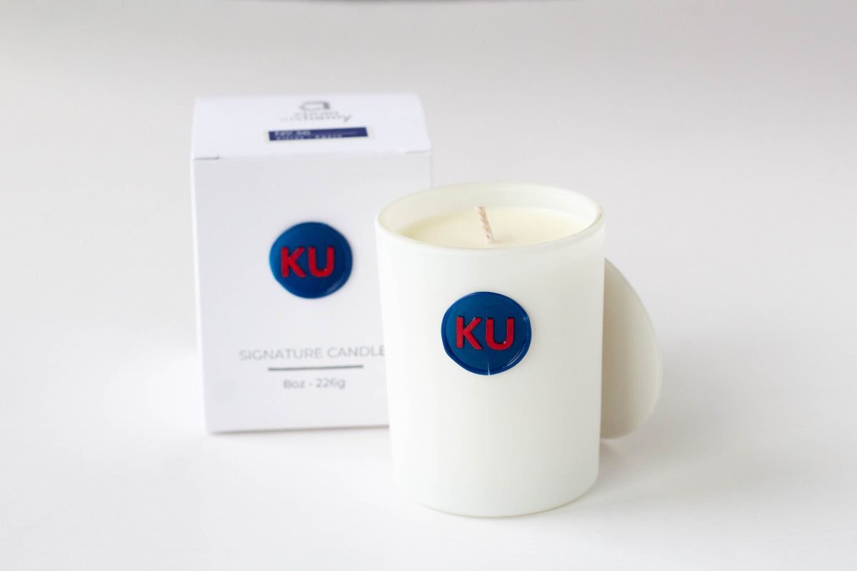A white ceramic candle jar with a blue circular 'KU' logo sits next to its white packaging box on a clean white background.