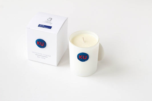 A white glass jar candle with cream-colored wax and a blue circular 'KU' logo, shown next to its white packaging box on a clean white background.