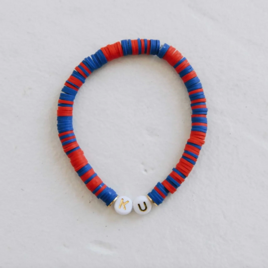A handmade beaded bracelet made of alternating red and blue paper discs, finished with two white round beads featuring the gold letters 'K' and 'U', displayed on a light textured background.