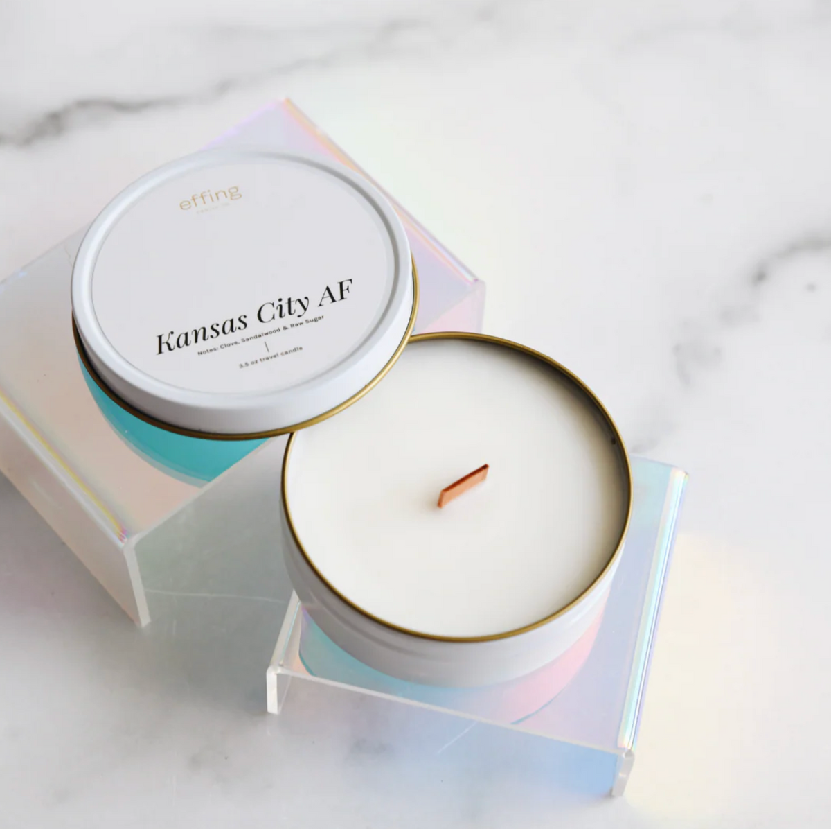 A white candle in a white travel tin sits on a reflective stand on a white marble surface. The candle label reads "Kansas City AF"