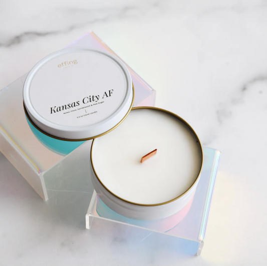 A white candle in a white travel tin sits on a reflective stand on a white marble surface. The candle label reads "Kansas City AF"