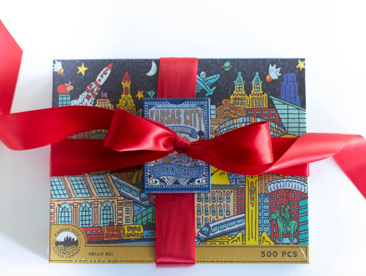 A boxed puzzle wrapped with a red ribbon and bow, featuring a colorful illustrated Kansas City themed cityscape on the lid. The front label reads 'Hello KC' and '500 PCS'.