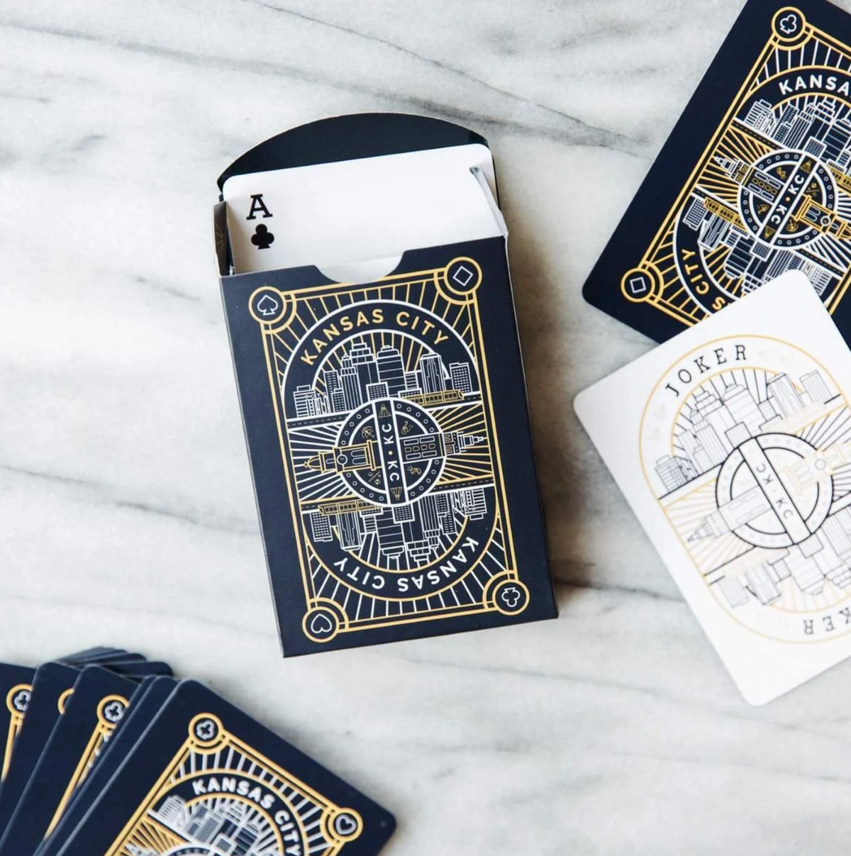 A styled flat-lay of a Kansas City playing card deck on a white marble surface, showing the black and gold illustrated packaging, a deck box with an Ace card, a single Joker card, and fanned cards.