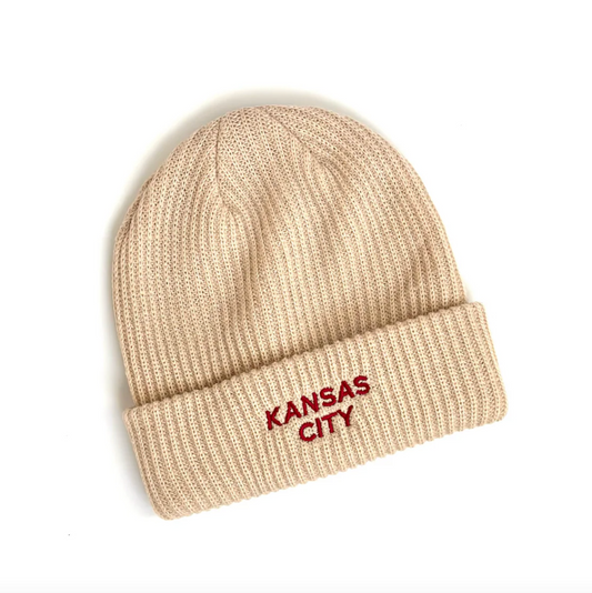 A beige ribbed knit beanie hat with a folded cuff, featuring the words 'KANSAS CITY' embroidered in red on the front cuff.