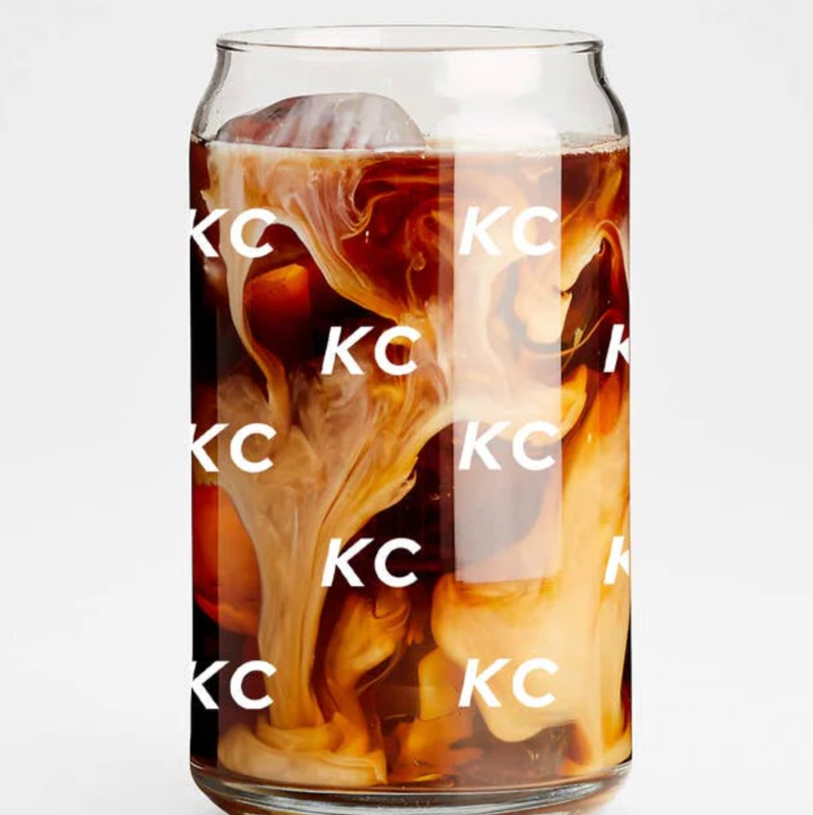 A clear glass can-shaped tumbler filled with iced coffee and milk swirls, covered in a repeating white 'KC' logo pattern, on a light gray background.