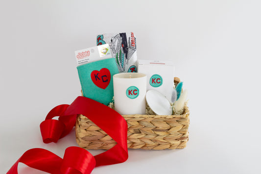 A woven gift basket filled with assorted items including a white ceramic candle with a teal KC logo, a teal drink sweater with a red heart and KC Current themed Kitchen Towel and a red ribbon bow, photographed on a clean white background.