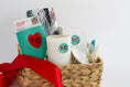 Load image into Gallery viewer, A woven gift basket filled with assorted items including a white ceramic candle with a teal KC logo, a teal drink sweater with a red heart and KC Current themed Kitchen Towel and a red ribbon bow, photographed on a clean white background.

