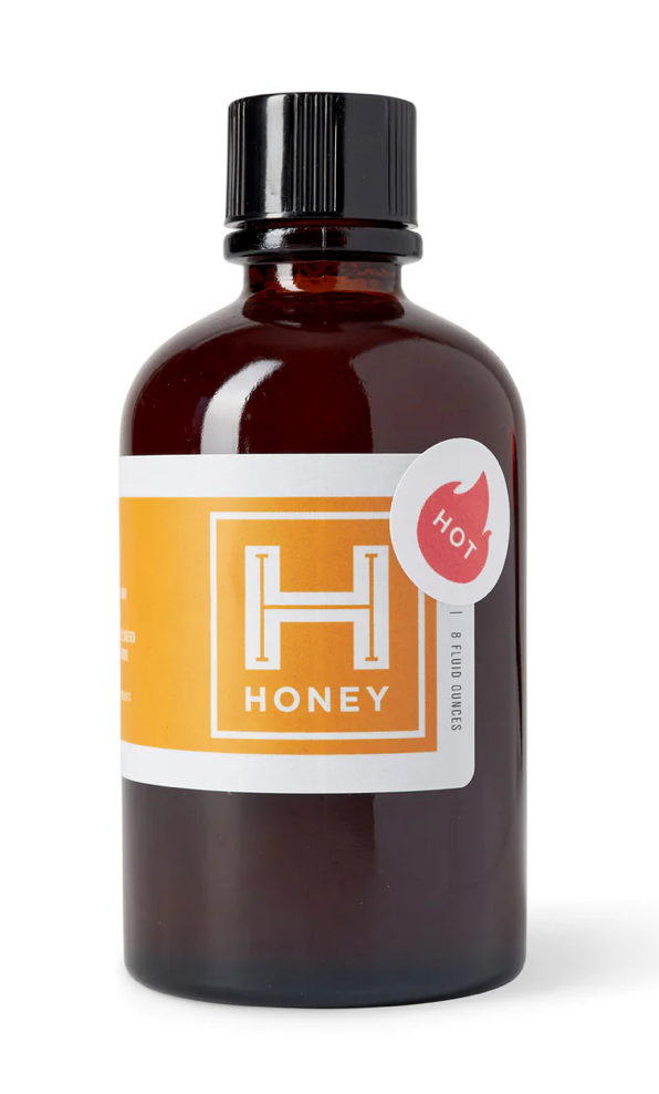 An amber glass bottle of hot honey with a black ribbed cap and a white and orange label featuring a large 'H' logo and a red 'HOT' badge, shown on a white background.