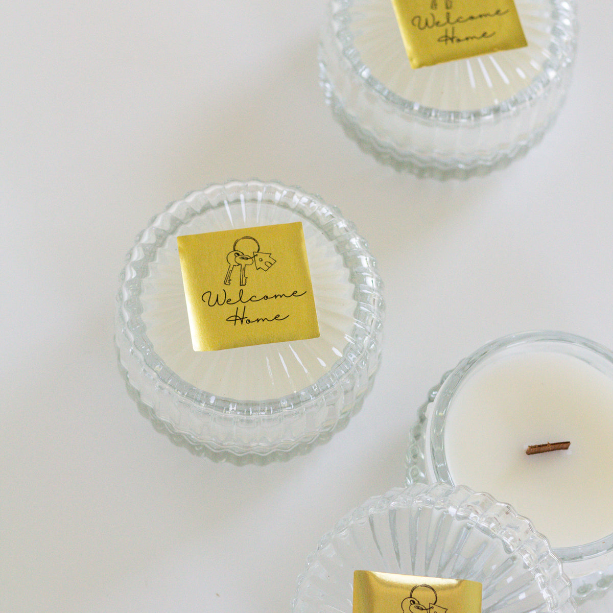 Three small glass jar candles with clear ribbed lids and white wax are arranged on a white background. Each lid has a gold square label reading 'Welcome Home' with a simple line drawing.