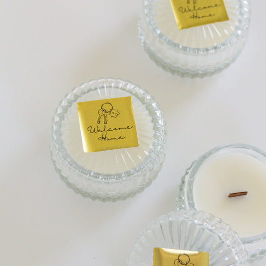 Three small glass jar candles with clear ribbed lids and white wax are arranged on a white background. Each lid has a gold square label reading 'Welcome Home' with a simple line drawing.