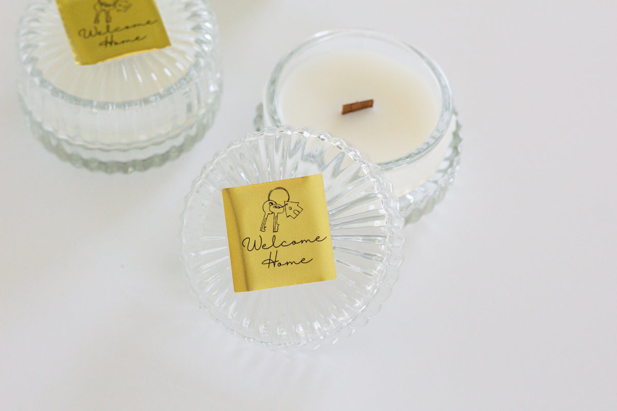 Two clear glass jar candles with gold 'Welcome Home' labels are arranged on a white surface. One candle is open, showing white wax and a small brown wood wick, while the other one is closed.